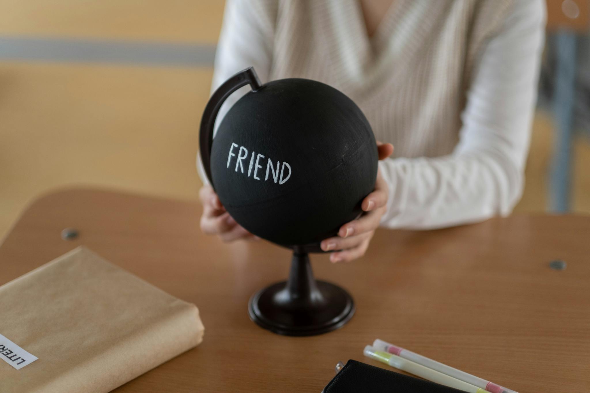 A close-up of a person holding a black globe inscribed with the word 'Friend' in a classroom setting.